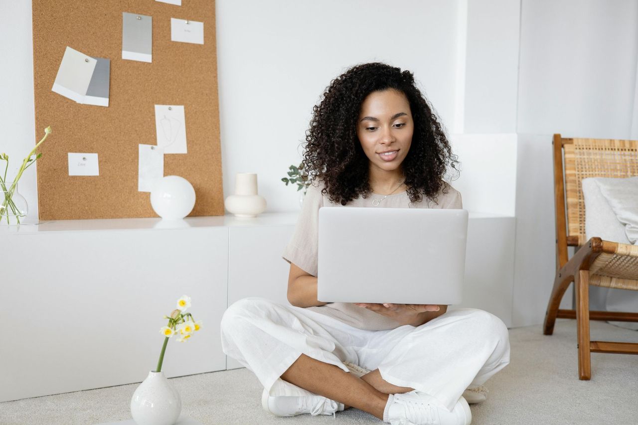 A person with dark curly hair sits cross-legged on the floor, using a laptop in a bright, minimalist home office.