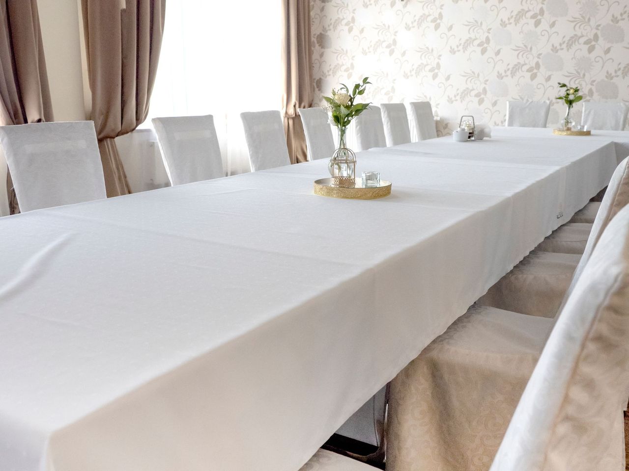 A long table covered with a white tablecloth, set with chairs and floral centerpieces in a bright, formal event room.